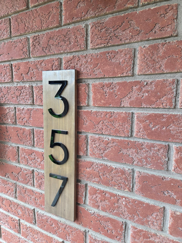 Modern vertical house number sign with 3D black acrylic digits "357" mounted on a light wood plaque against a red brick wall.