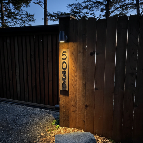 A vertical modern acrylic address sign with house number 5036, illuminated at night by a warm overhead light on a wooden fence post.